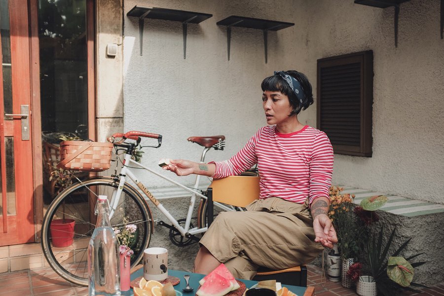 Ade mengenakan kaus garis-garis merah, sedang duduk di hadapan meja yang terdapat potongan buah dan secangkir minuman.