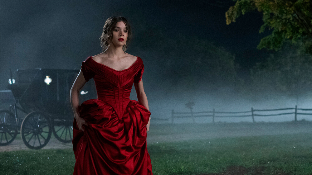 Hailee Steinfeld mengenakan gaun merah, berdiri di depan kereta kuda, pada setting malam hari.