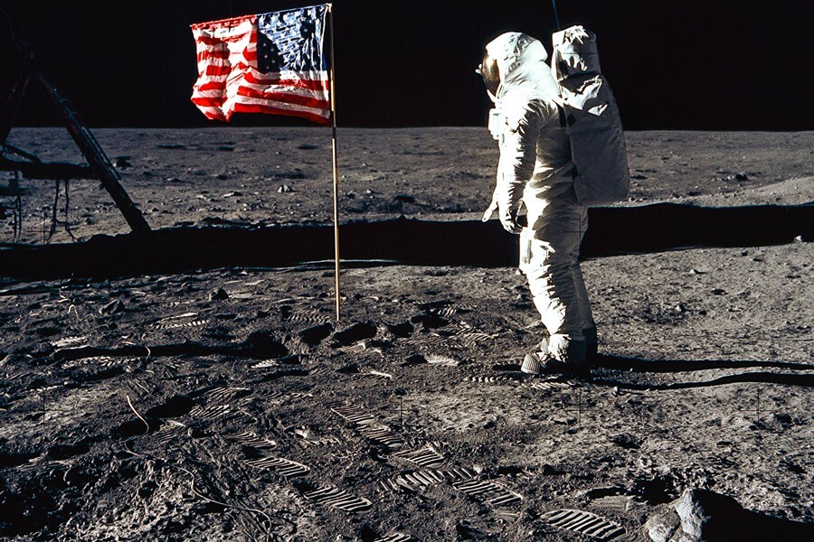 Seorang astronot berpakaian putih di luar angkasa, terlihat seperti baru saja menancapkan bendera Amerika Serikat.