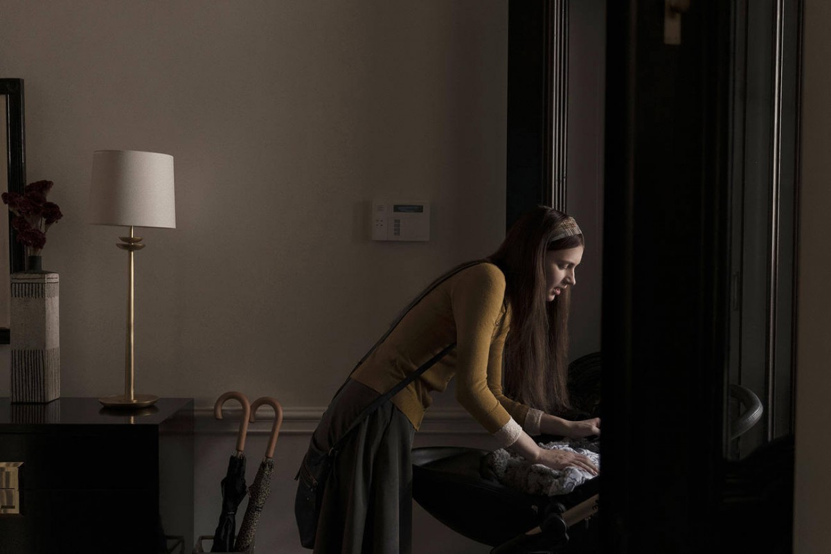 Seorang wanita berpakaian kuning terlihat sedang menidurkan bayi, ada dua puah bayung dan lampu di belakangnya.