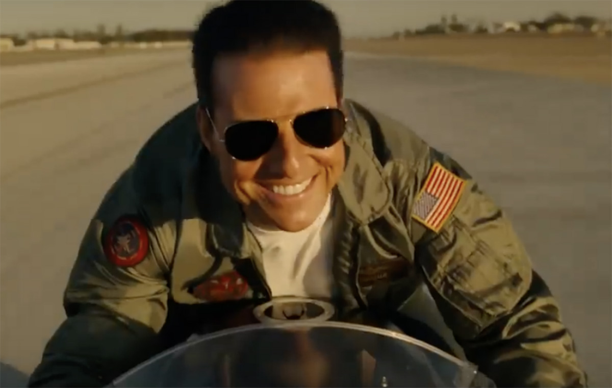 Foto close-up Tom Cruise sedang mengendarai sepeda motor sambil tersenyum.