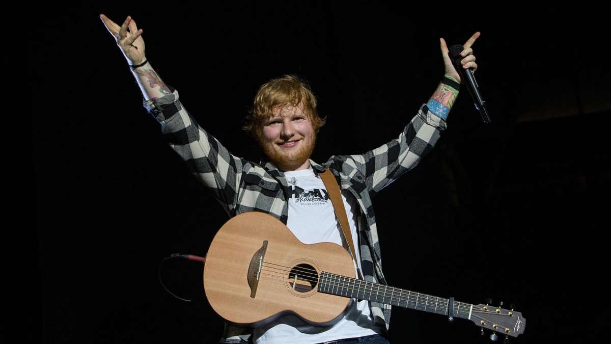 Ed Sheeran mengenakan kaus putih dan kemeja kotak-kotak, mengalungkan gitar, berdiri di atas panggung.