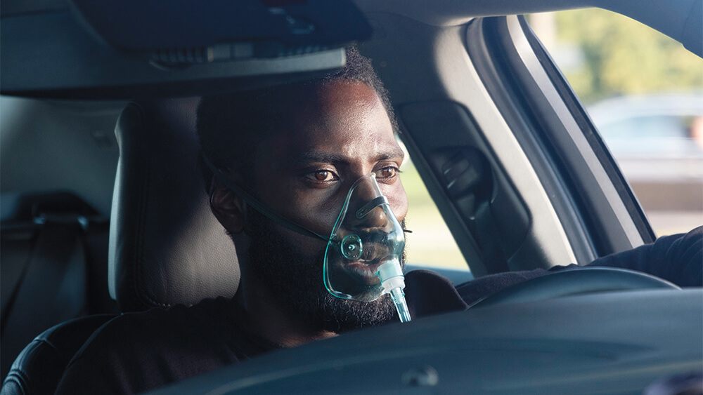 Salah satu adegan film Tenet di mana John David Washington terlihat menggunakan masker dan terdiam di dalam sebuah mobil