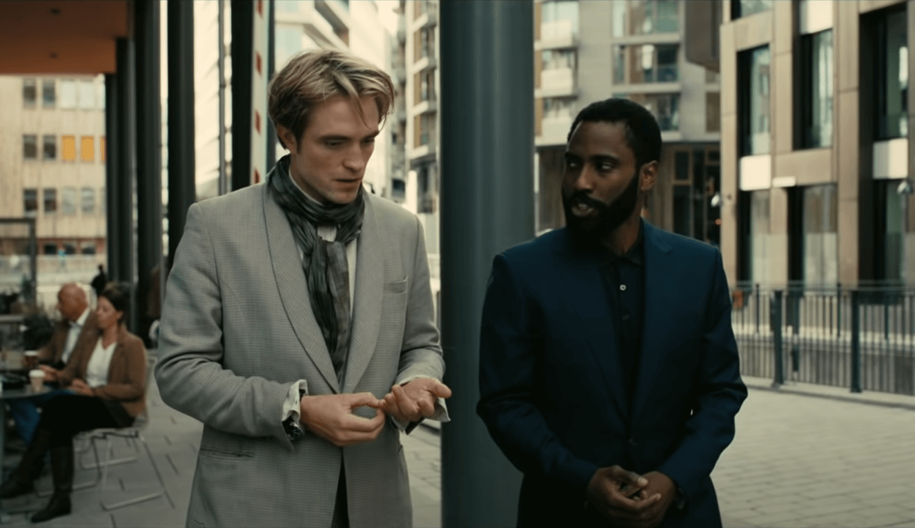 Robert Pattinson dan John David Washington sedang berjalan dalam balutan jas bewarna abu-abu dan navy blue di salah satu scene Tenet